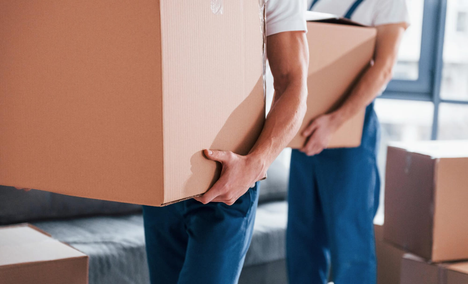 two young croped movers with blue uniform working indoors and holding one moving box each